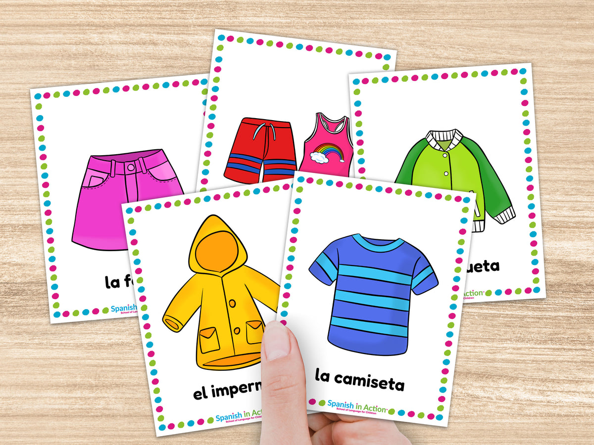 A hand holds a fan of Spanish clothing flashcard illustrations on a light wood surface. The cards show colorful drawings of garments such as pink skirt, red striped swimsuit, a pink swimsuit, a green jacket, and a blue striped shirt, with Spanish labels and a multicolored polka-dot border.